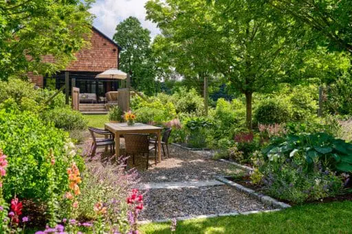 Boston-area garden designed with a gravel dining terrace, layered perennial plantings, mature trees, and a welcoming outdoor seating area.