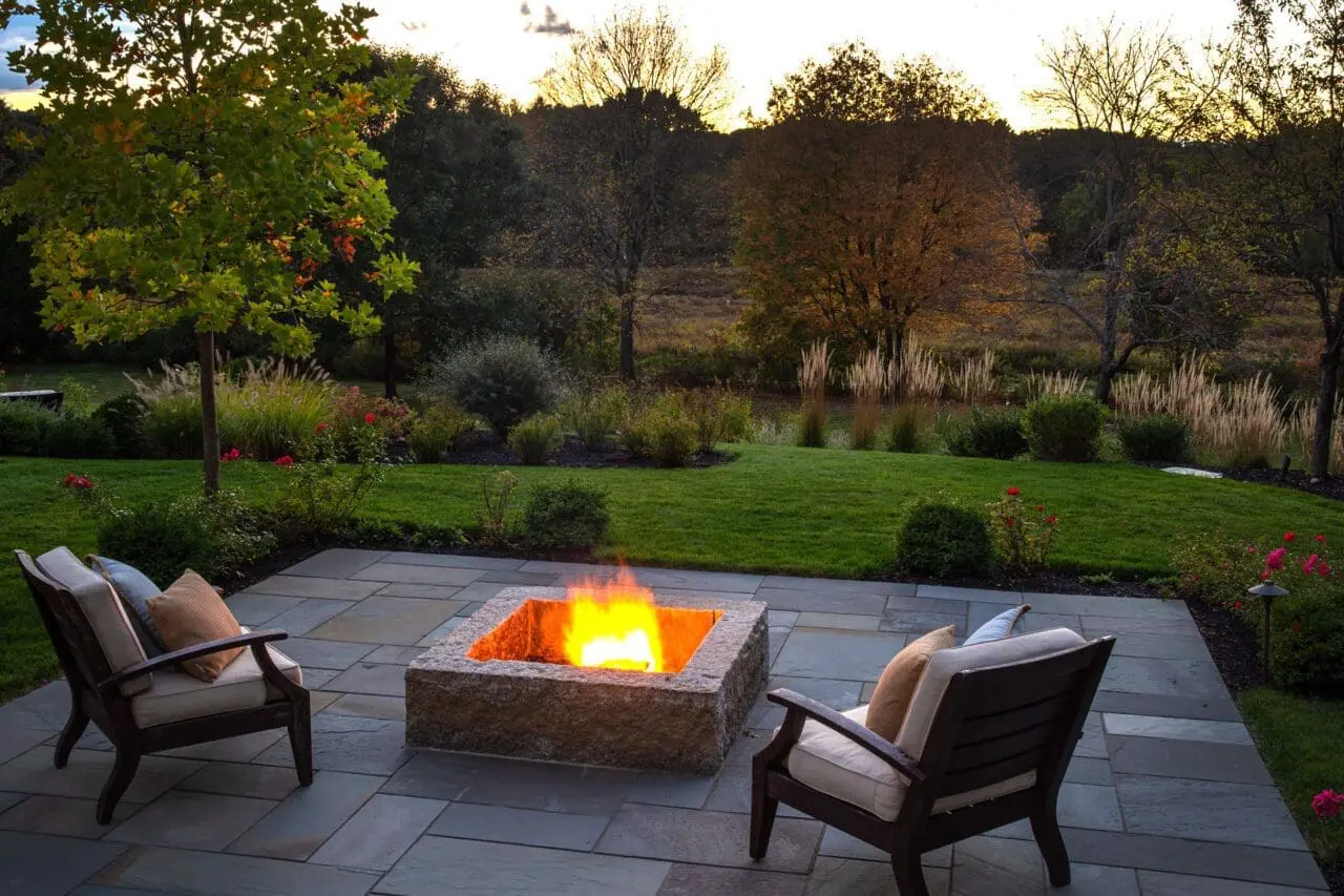 A stone patio and fire pit overlook a lush garden at dusk, creating an inviting outdoor retreat designed for comfortable evenings outside.