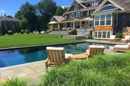 A Duxbury backyard pairs a rectangular pool and stone terrace with a broad lawn, ornamental grasses, and relaxed seating beside a shingle-style home.