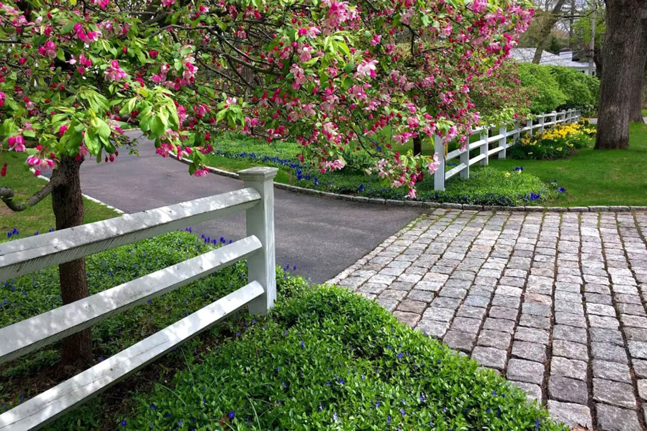 A cobblestone driveway framed by a flowering tree and white fence delivers a charming spring entry, combining texture, durability, and traditional detailing.