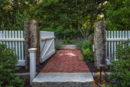 Lincoln, MA - A white garden gate framed by stone posts creates a graceful transition from walkway to garden, blending structure with softness.