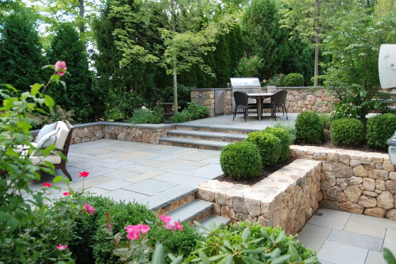 Stone retaining walls surround the bluestone patio and outdoor kitchen, with shrubs and hedges providing privacy, wind protection, and a lush backdrop.