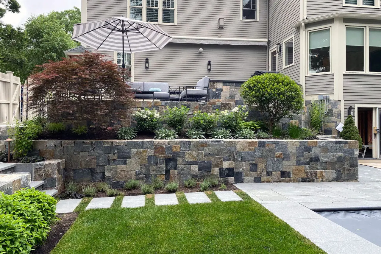 Belmont, MA - Granite block steps are integrated into a sloped retaining wall, with adjacent shrubs stabilizing the grade and visually tying the stonework into the landscape.