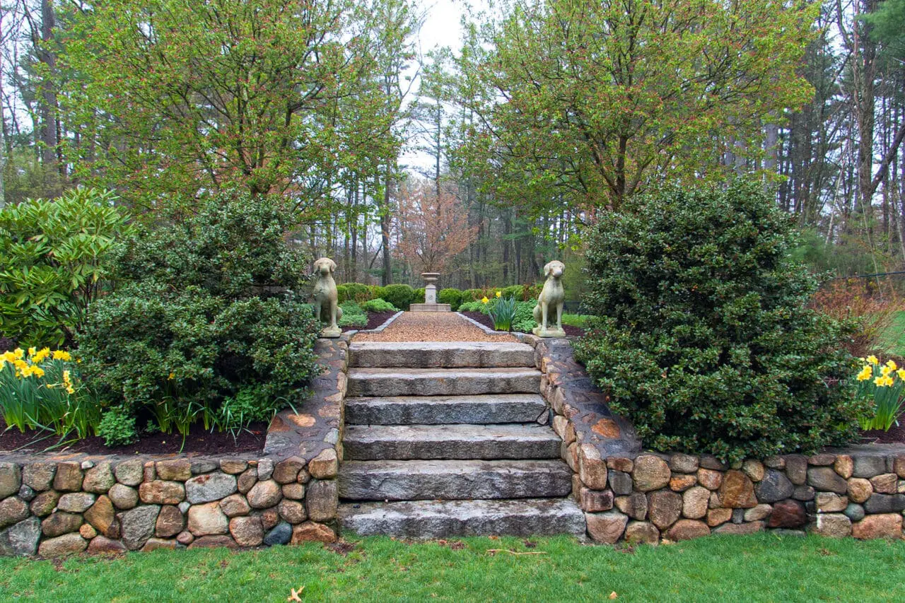 Granite steps and fieldstone walls establish a strong formal axis, using symmetry and scale to organize planting beds and clipped hedges along the garden’s main sightline.
