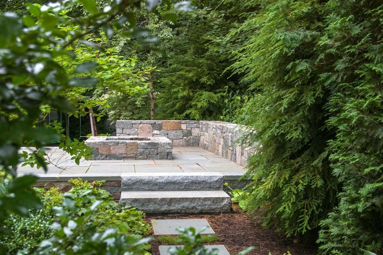 Wellesley, MA - A secluded stone fire pit patio offers a quiet gathering place tucked into mature evergreens.