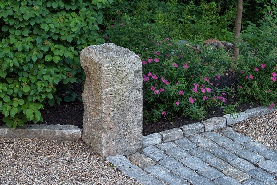 A reclaimed granite post and cobblestone edging add age and texture to a gravel path bordered by lush flowering plantings.