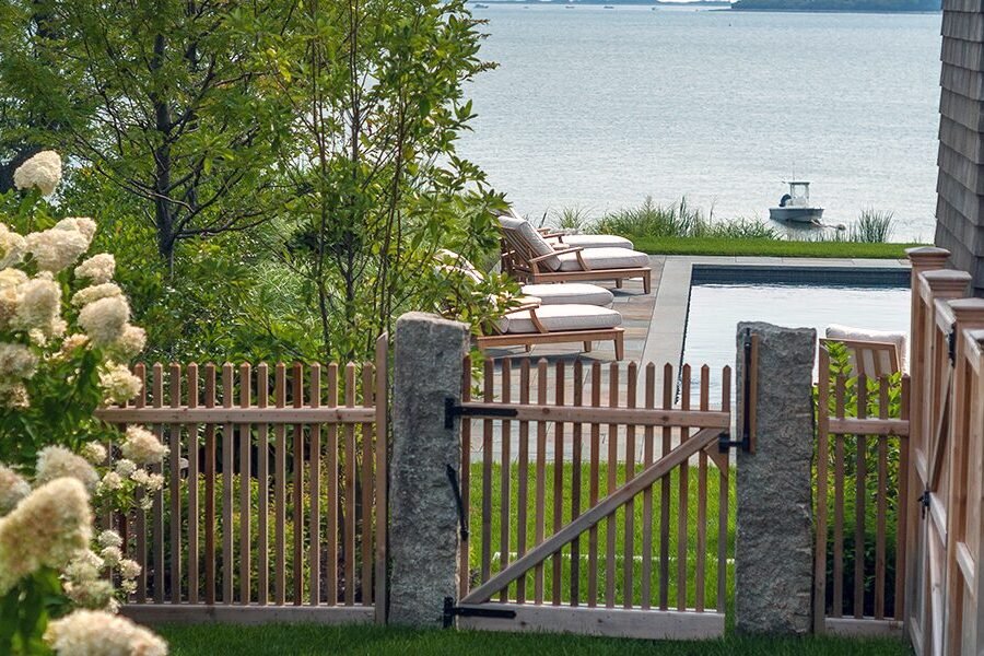 Reclaimed granite gateposts frame a simple wood gate, opening to a waterfront pool and relaxed coastal landscape.