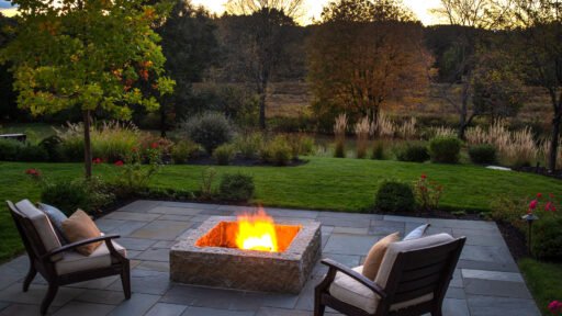 A stone patio and fire pit overlook a lush garden at dusk, creating an inviting outdoor retreat designed for comfortable evenings outside.