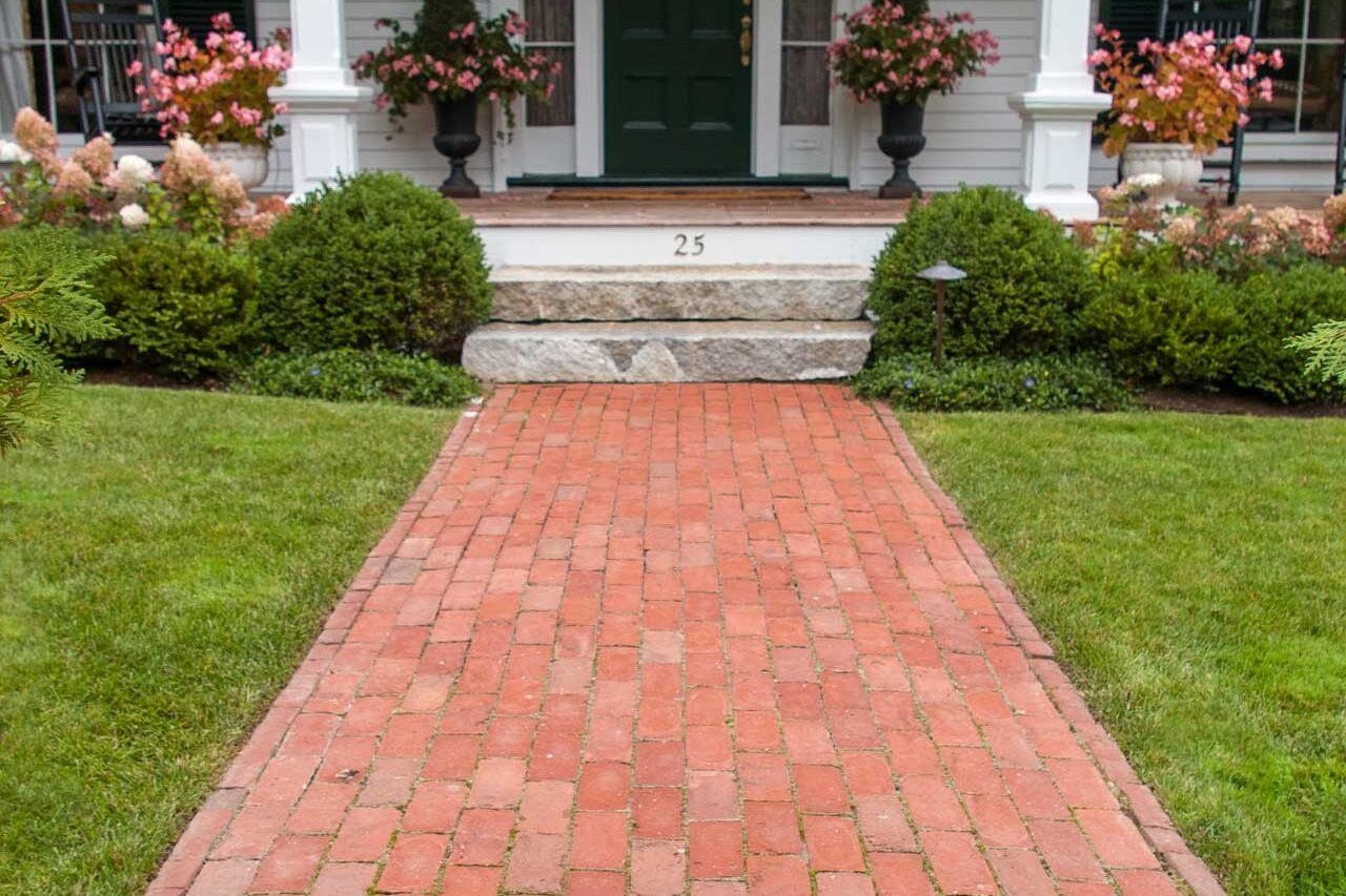 Belmont, MA - Classic clay brick walkway and antique granite steps lead the way to a sprawling front porch accented by colorful planters.