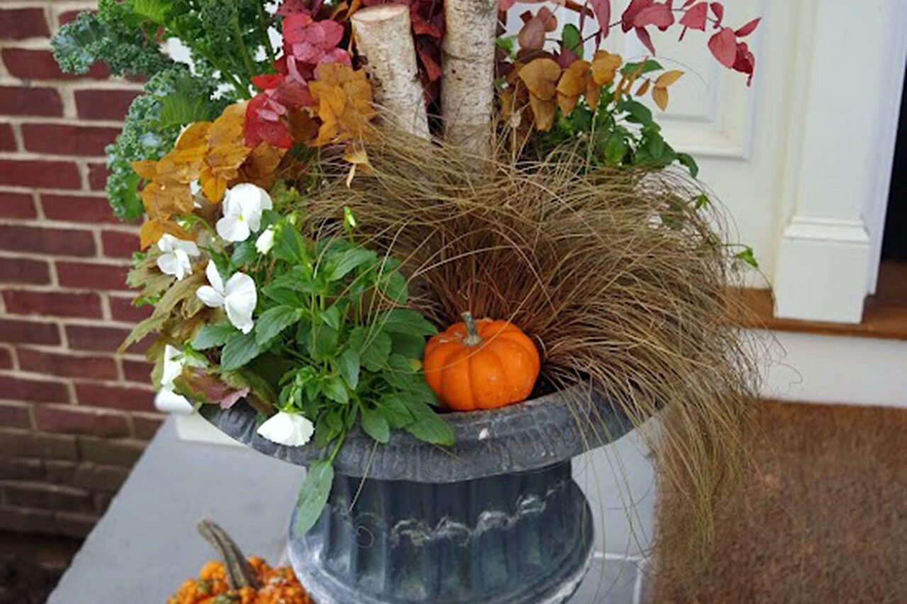 A black urn at the porch entry filled with birch logs, fall foliage, and pumpkins creates a timeless autumn display, blending natural materials and classic styling for flexible, season-long curb appeal.