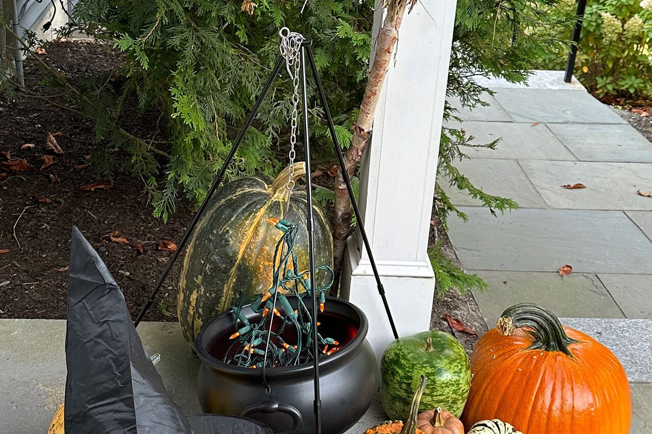 A cauldron planter paired with gourds and a witch hat adds a whimsical Halloween accent along the front walk, balancing spooky fun with landscape design details.