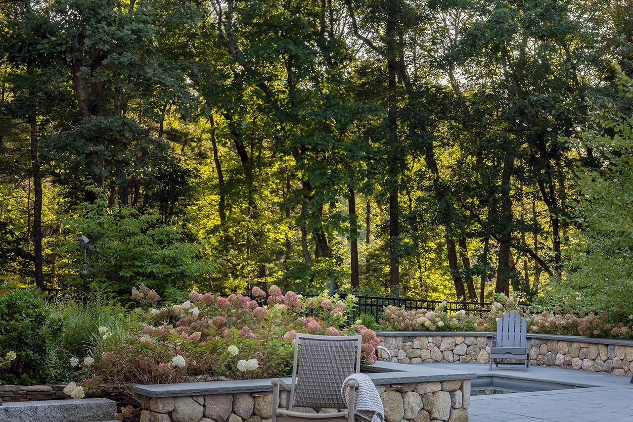 Surrounded by trees, this square stone fire pit brings warmth and definition to a bluestone patio, creating an inviting destination that feels tucked into the woodland landscape.