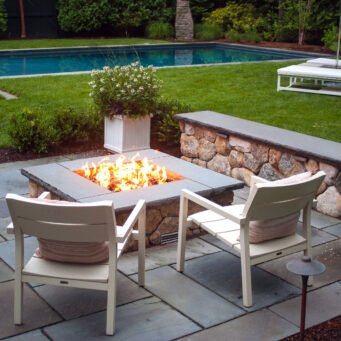 A square stone fire pit anchors this bluestone terrace beside the pool, offering clean lines, durable materials, and a central gathering point that enhances both day and evening use.