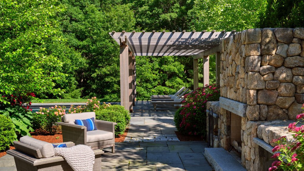 A stone outdoor fireplace beneath a pergola defines this lounge patio, adding shade, structure, and warmth while creating a year-round destination for relaxed seating and conversation.