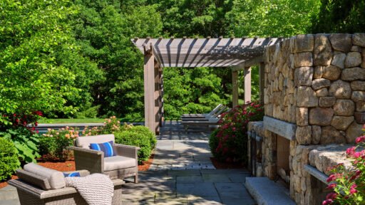 A stone outdoor fireplace beneath a pergola defines this lounge patio, adding shade, structure, and warmth while creating a year-round destination for relaxed seating and conversation.