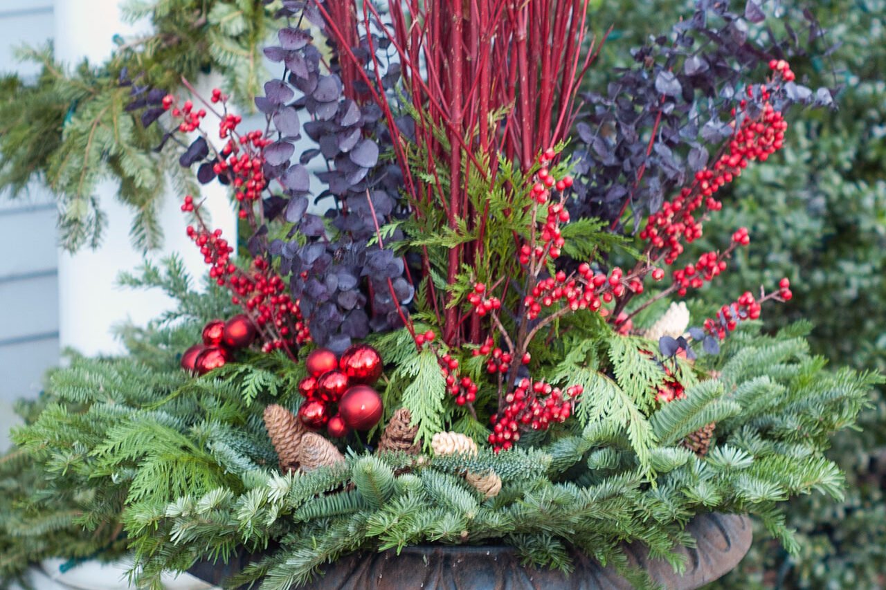 A cast iron urn filled with evergreen boughs and red twig accents creates a festive winter porch display with lasting structure.