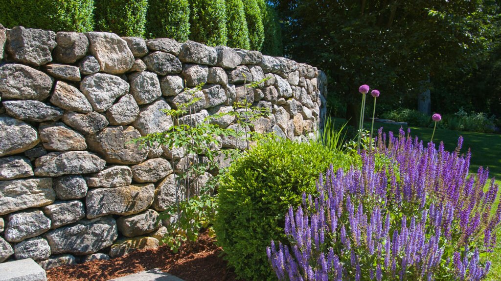 A dry-laid fieldstone retaining wall defines the garden edge, using irregular, hand-set stone to manage a subtle grade change while creating a low, natural boundary with a relaxed, timeless character.