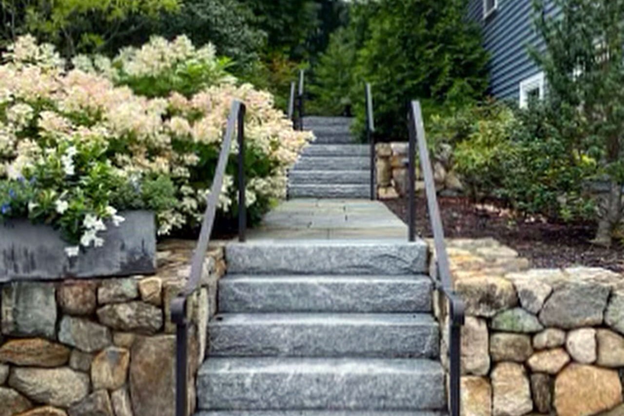 Granite front steps and flanking fieldstone walls create a formal entry, balanced by shrubs and hedges that soften the stone and reinforce symmetry.