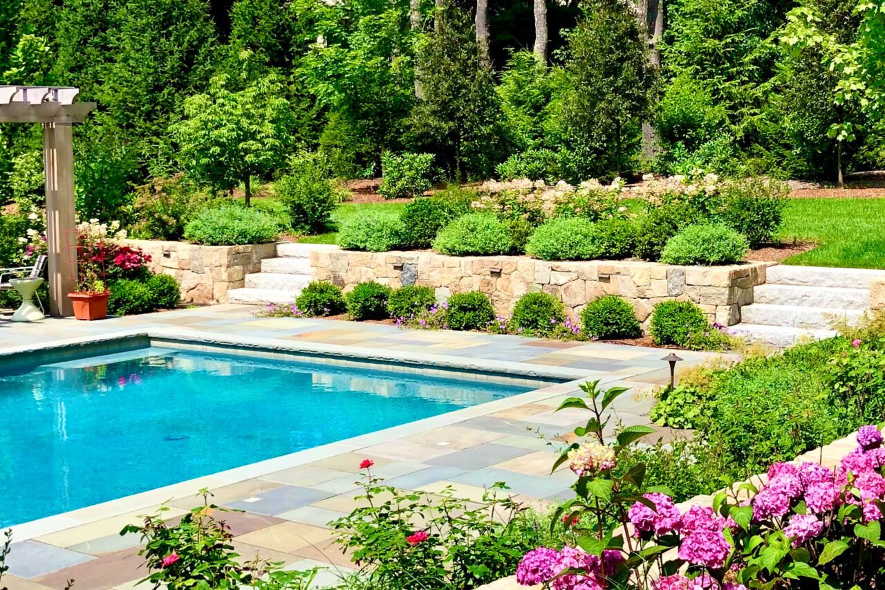 A stone retaining wall and bluestone steps define circulation around the pool terrace, balancing durable materials with surrounding shrubs that provide enclosure and visual relief from the hardscape.