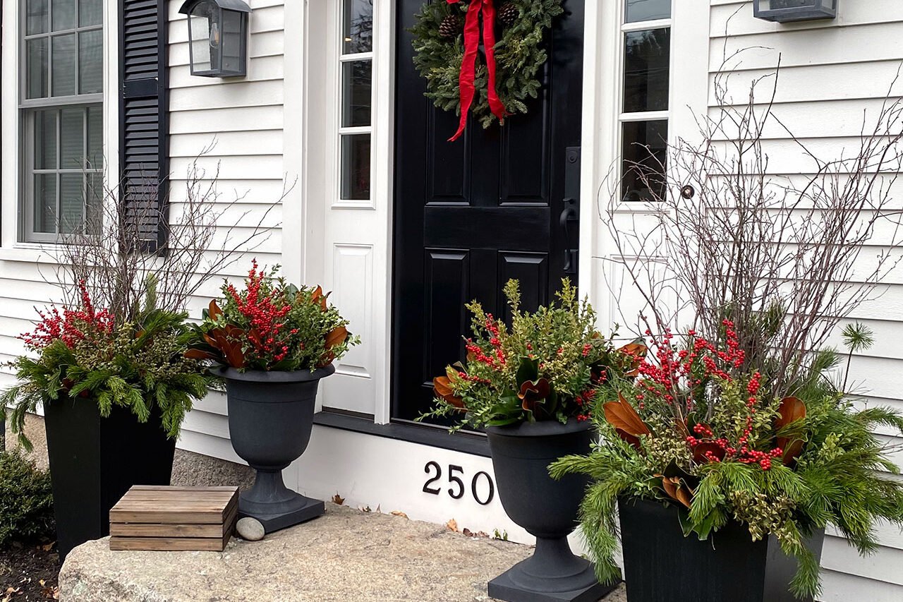 New England entryway with a black front door decorated with a holiday wreath and winter planters filled with seasonal greenery.