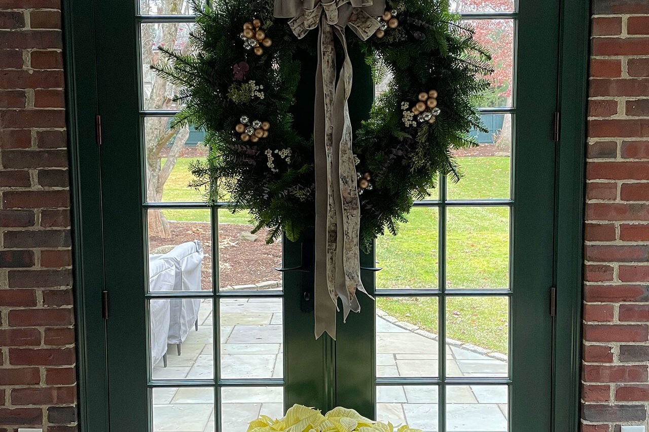 Holiday wreath with a soft taupe bow on dark green French doors, framed by evergreen garland and brick walls, with a white poinsettia below.